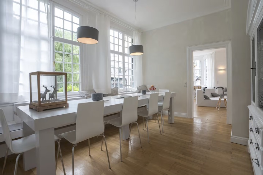 Ansicht des hellen Esszimmers mit Platz für 12 Personen, große Fenster zum Garten, im Hintergrund führt die offene Tür zum Wohnzimmer